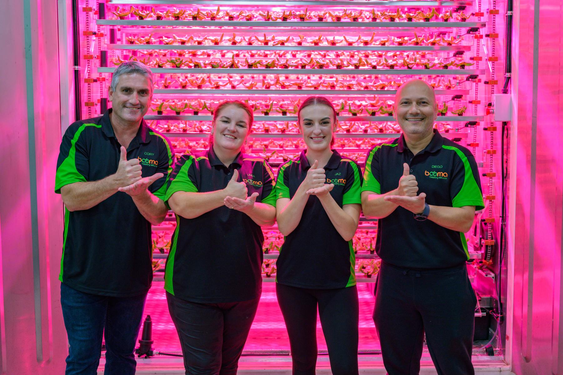 Four colleagues with their thumbs up, smiling at camera in front of converted shipping container lit pink and filled with racks of seedlings