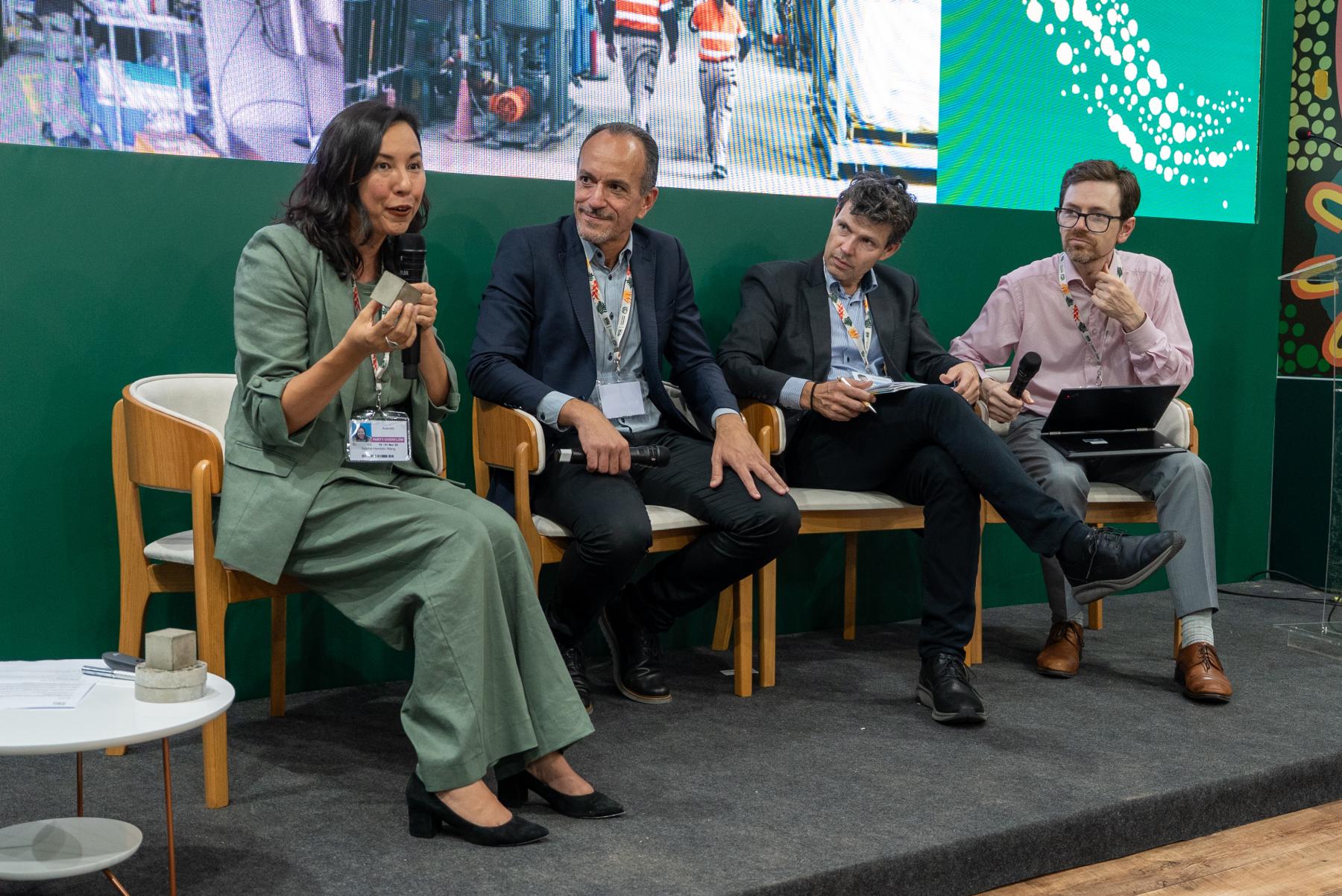 Four people sitting on a panel, three men watching a woman holding a microphone