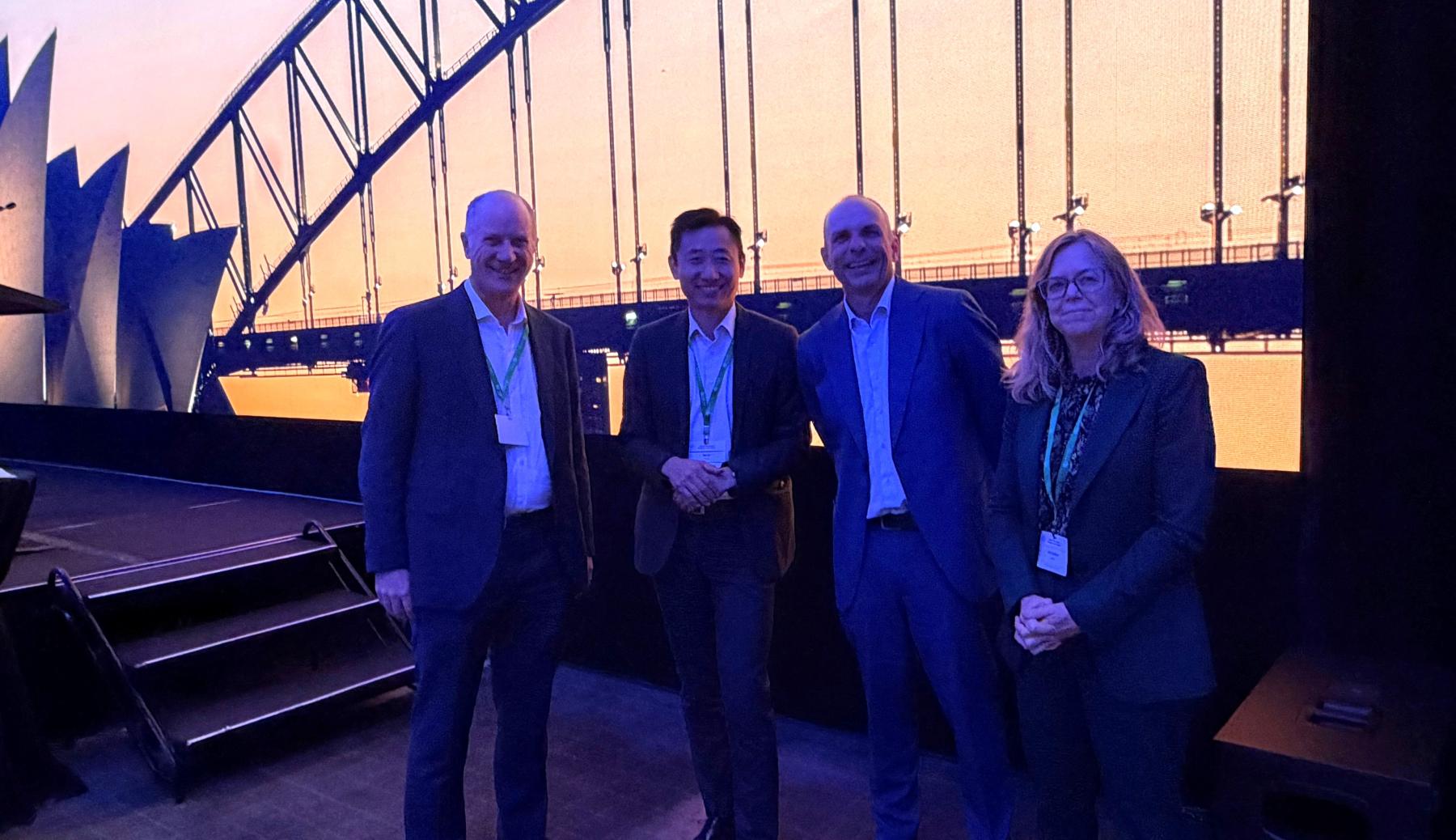Four people stand in front of a stage with a backdrop of Sydney harbour.