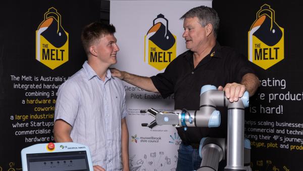 An older man holding a young man's shoulder and a mechanical arm in front of banners for The Melt