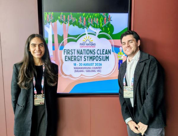 Two NZEA staff stand in front of a sign for the First Nations Clean Energy Symposium