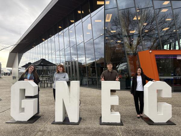Four people outside the conference venue standing in front of the letters G N E C