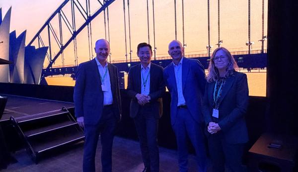 Four people stand in front of a stage with a backdrop of Sydney harbour.